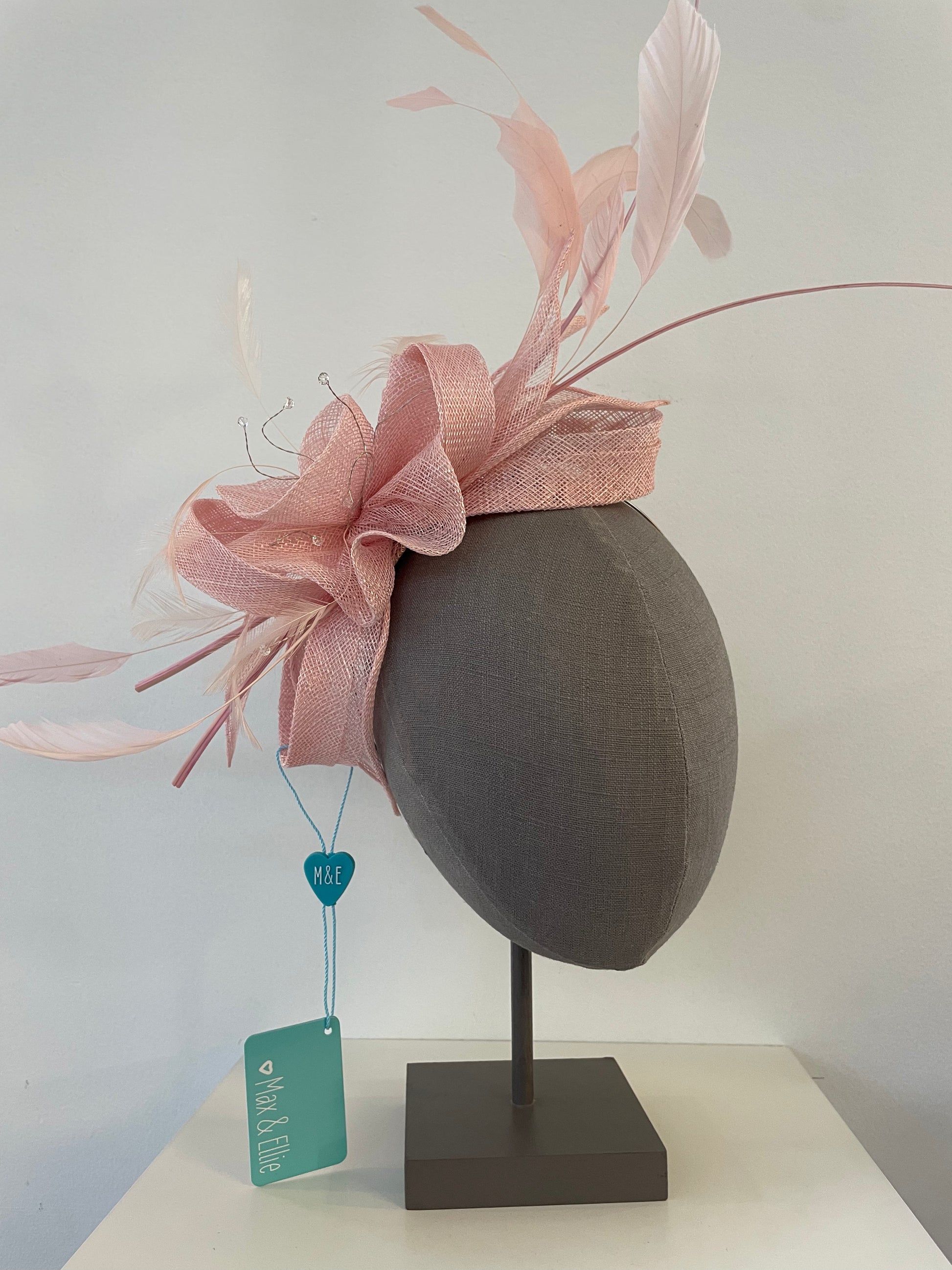 A light pink fascinator hat with matching pink mesh ribbon and feather accents, displayed on a dark gray fabric mannequin head on a stand.
